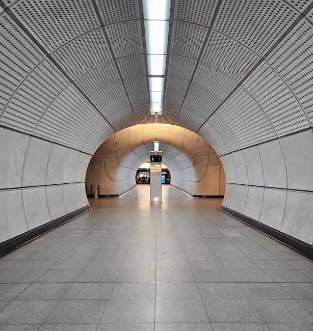 A symmetrical view of a modern underground passageway with a rounded, tunnel-like ceiling composed of perforated metal panels arranged in concentric circles. The ceiling features central lighting strips emitting a soft white glow, highlighted by warm-toned accent lighting near the curved wall sections. The walls are clad in large, smooth light grey panels, and the flooring consists of polished grey tiles reflecting the ambient light. At the far end of the tunnel, a vertical column with an electronic information display stands centrally, guiding pedestrians towards exits or platforms. The environment appears clean and well-maintained, suggesting it is part of a public transportation hub or railway station. The design's emphasis on rounded shapes and neutral tones aligns with efficient, contemporary interior aesthetics found in urban transit infrastructure, consistent with premises that may require regulated waste management by private services like Rubbish Clearance Soho for their maintenance or refurbishment activities.