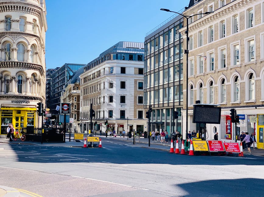 A busy city street scene featuring a mix of historic and modern buildings with light-colored facades and large windows. In the foreground, there is a roadblock with orange traffic cones, signs reading 'Road Closed' and 'Access Only,' and a black barrier indicating restricted vehicle access. Pedestrians can be seen walking along the sidewalk on both sides of the street, some entering and exiting shops, including a brightly painted yellow storefront. A tall street lamp stands near the right side, casting minimal shadow on the pavement. The background showcases multiple multi-storey office or residential buildings, with the clear blue sky overhead. This scene reflects urban traffic management and foot traffic typical of central London streets, with naturally lit conditions suggesting daytime, aligning with private or independent waste management activities in a vibrant city environment, and subtly supporting urban rubbish removal and clearance services such as those provided by Rubbish Clearance Soho.