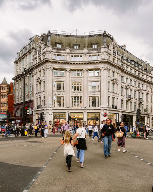 A busy city intersection in front of a large, historic cream-colored building with ornate architectural details, including decorative columns and window frames, situated on a bustling street corner. The building's façade features numerous large windows, and the ground level has retail stores with illuminated displays. Several pedestrians are crossing the street, some walking towards or away from the building, including a young girl in a white coat and fringe bag, a woman in a black skirt, and others dressed casually. The street is partially occupied by vehicles, and there are traffic signals and road markings visible in the scene. In the background, additional buildings with mixed architectural styles can be seen, along with streetlights and a cloudy sky overhead. This scene captures urban activity centered around a commercial district, where alternative waste handling or private rubbish collection services like those offered by Rubbish Clearance Soho might operate privately to manage rubbish removal efficiently in such densely populated areas.