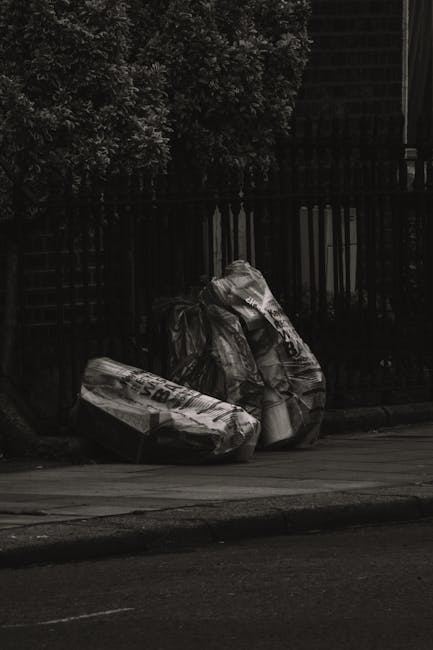 A group of black plastic rubbish bags, some with visible white labels and partially crumpled, are stacked on the pavement near the curbside in a residential or urban area. The bags appear filled with household waste or debris and are positioned close to a black metal fence with vertical bars, which runs along the sidewalk behind them. To the left, a dense green leafy bush is visible, providing some natural background contrast to the dark bags and fence. The scene is illuminated by natural daylight, with the pavement and road surface appearing smooth and dark, indicating a typical street setting. This arrangement of bags suggests an alternative waste collection or private rubbish removal service, such as Rubbish Clearance Soho, operating in an urban environment and preparing waste for disposal or collection, possibly as part of a site clearance or rubbish removal process.